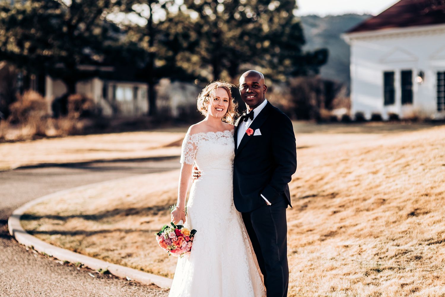 Bride in lace gown and groom in tuxedo pose together on sun-lit grounds.