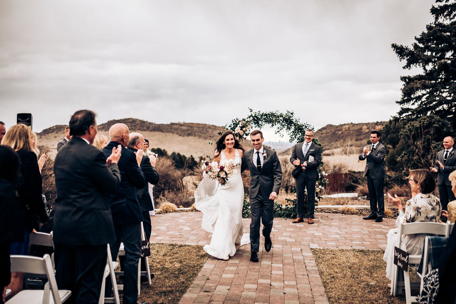 Wedding ceremony recessional on stone pathway with guests applauding and mountains in view.