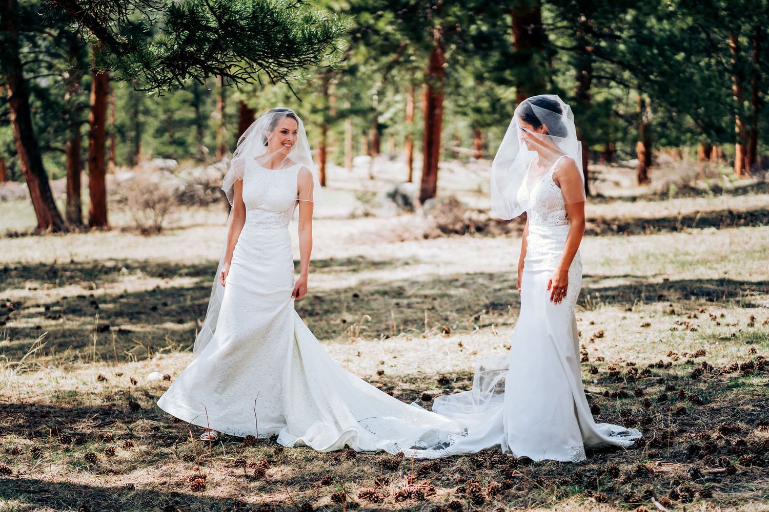 Two brides in white wedding dresses stand together in a sunlit forest setting.
