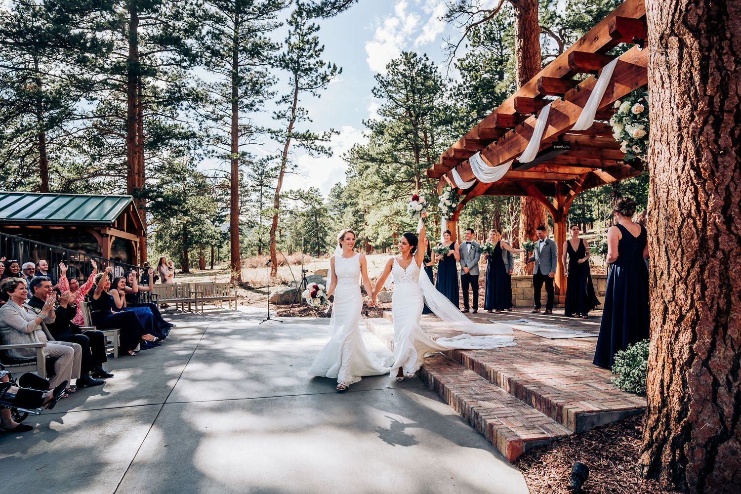 A wedding ceremony takes place on a rustic outdoor deck surrounded by tall pine trees in a mountain setting.