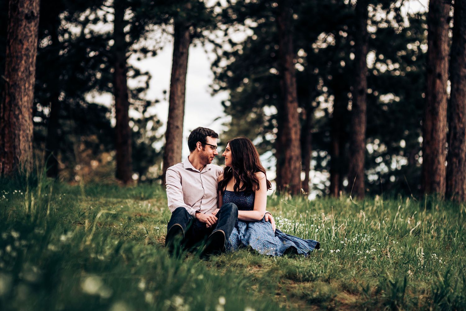 A couple sits together on a grassy hillside surrounded by tall pine trees.