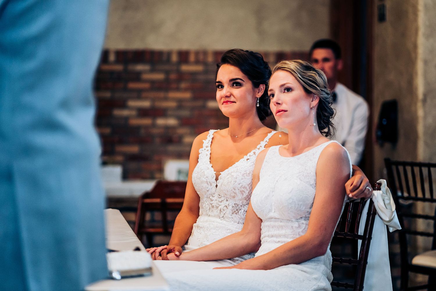 Two people in elegant white dresses seated at wedding reception table listening attentively.
