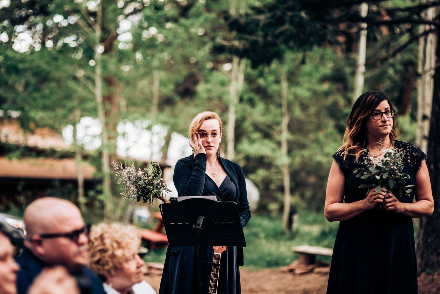 Wedding attendants in black dresses hold bouquets during an outdoor forest ceremony.