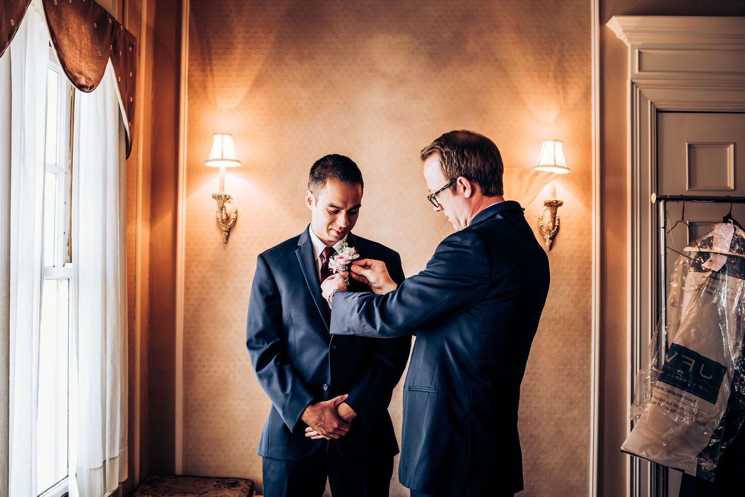Two people in suits helping with formal attire adjustments near window.