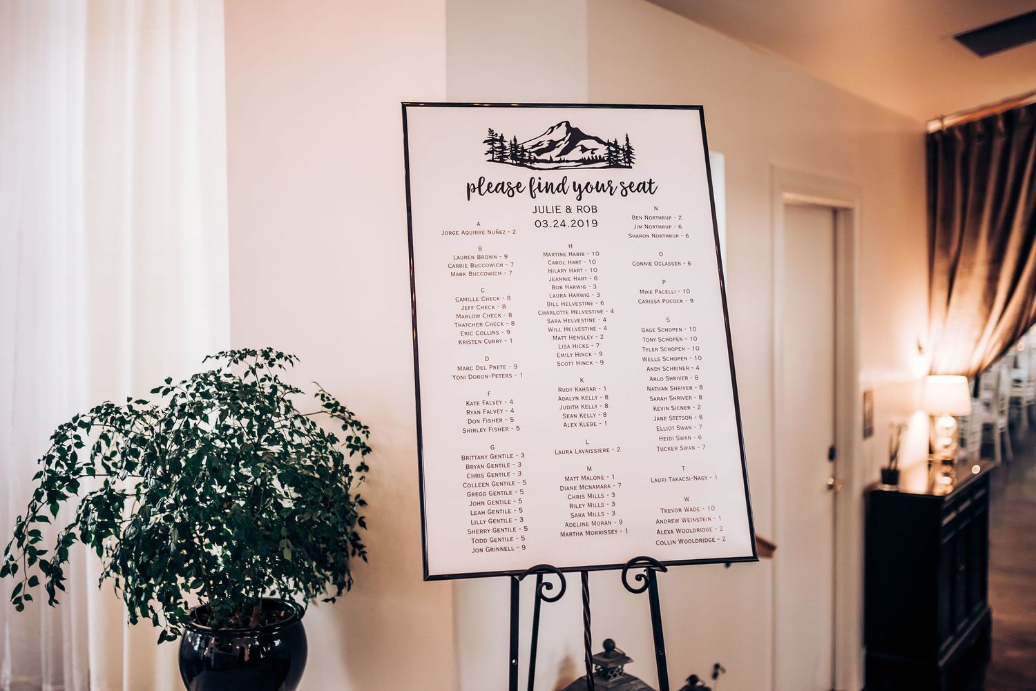 Wedding seating chart displayed on easel stand with potted plant decoration.