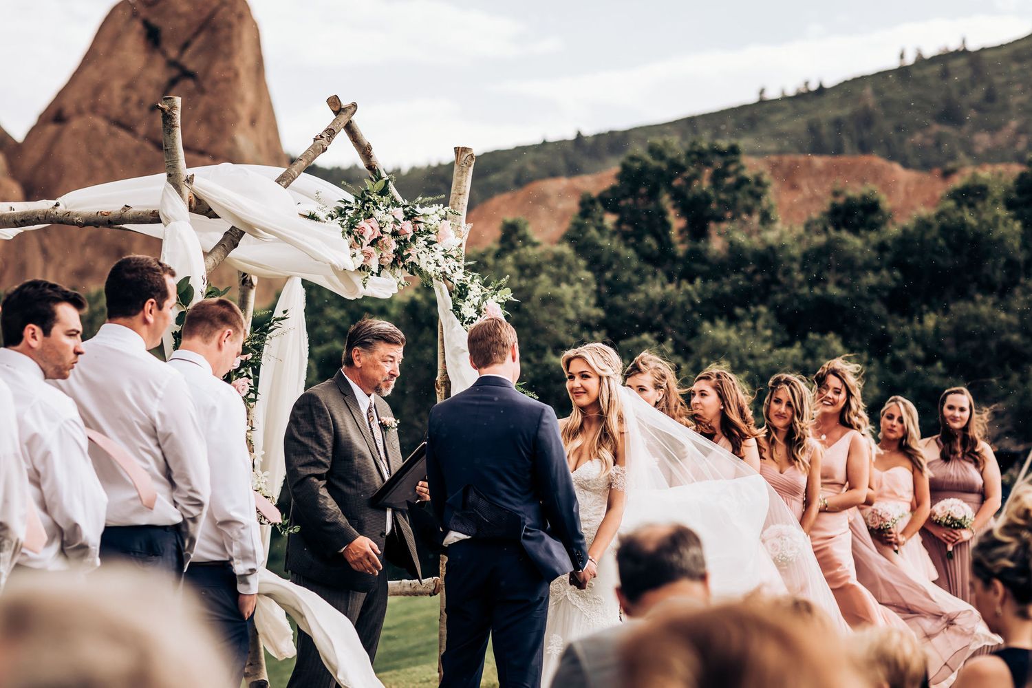 Wedding ceremony takes place under floral arch with mountain views and pink-clad attendants.