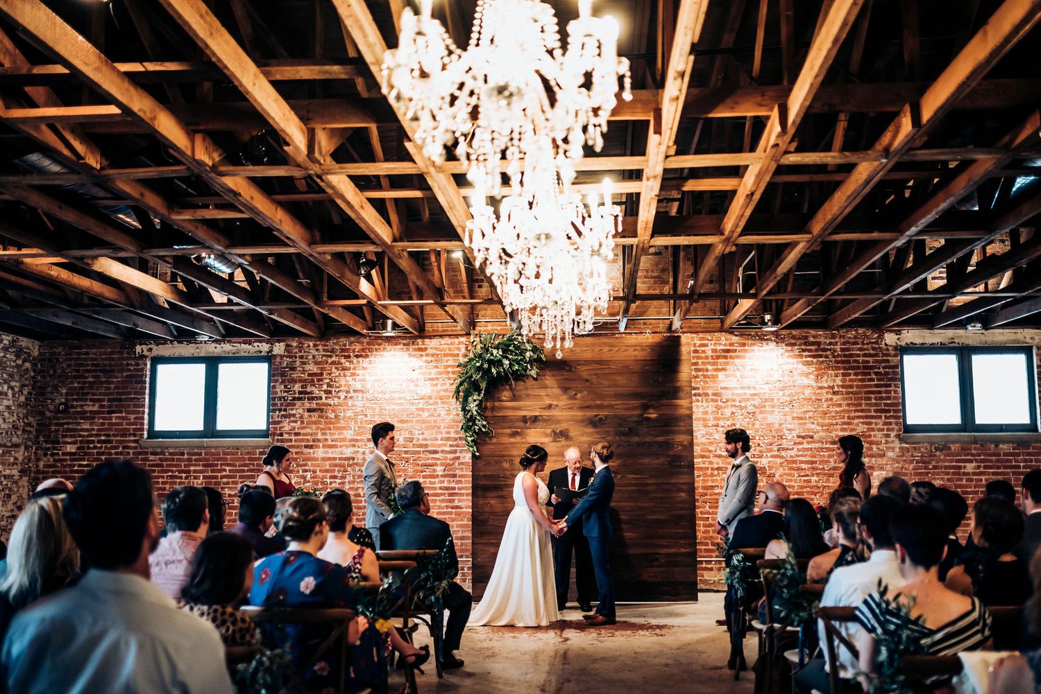 Wedding ceremony under crystal chandeliers in rustic venue with exposed brick and wooden beams.