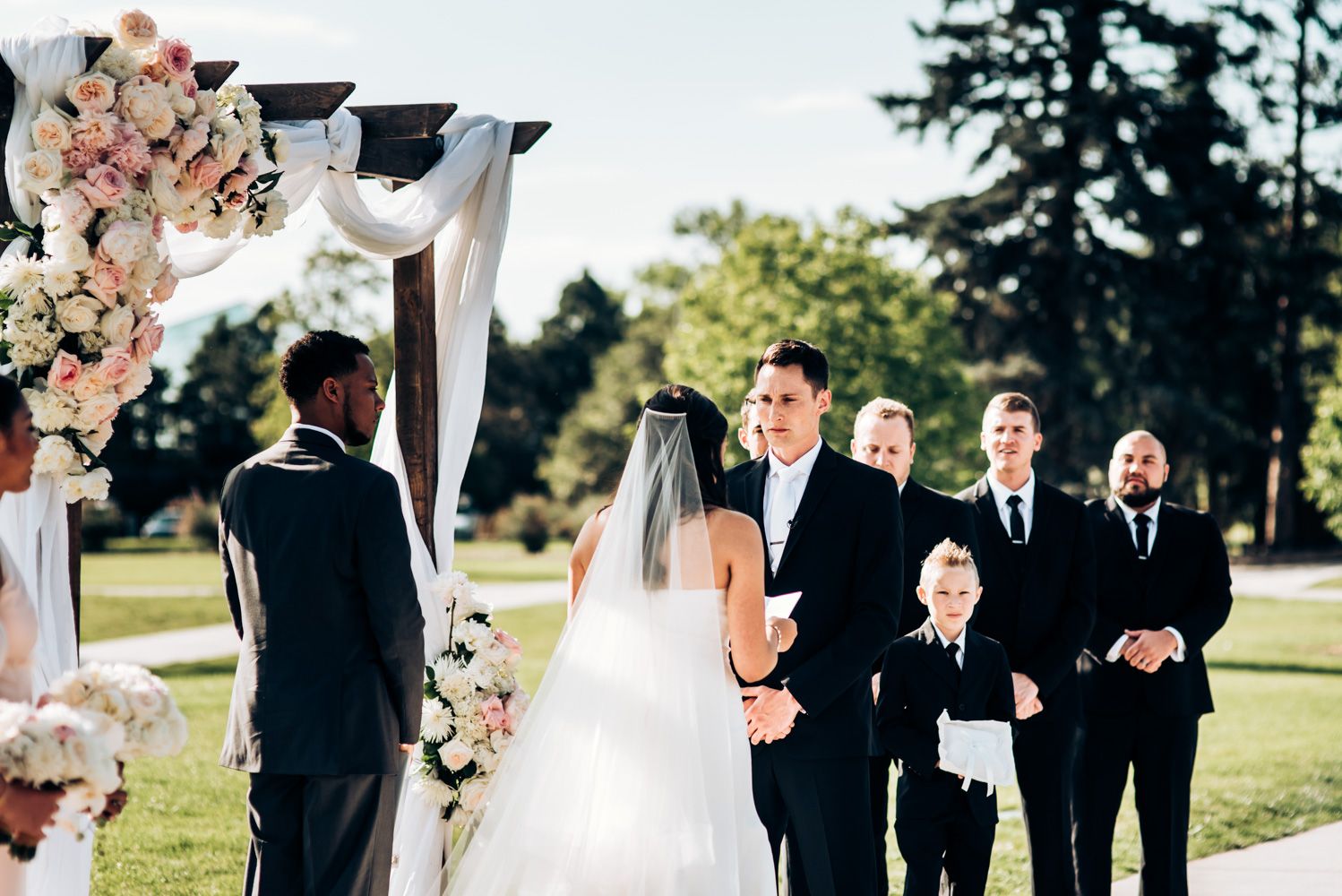 Wedding ceremony at an outdoor venue with floral arch and white fabric decorations in natural setting.