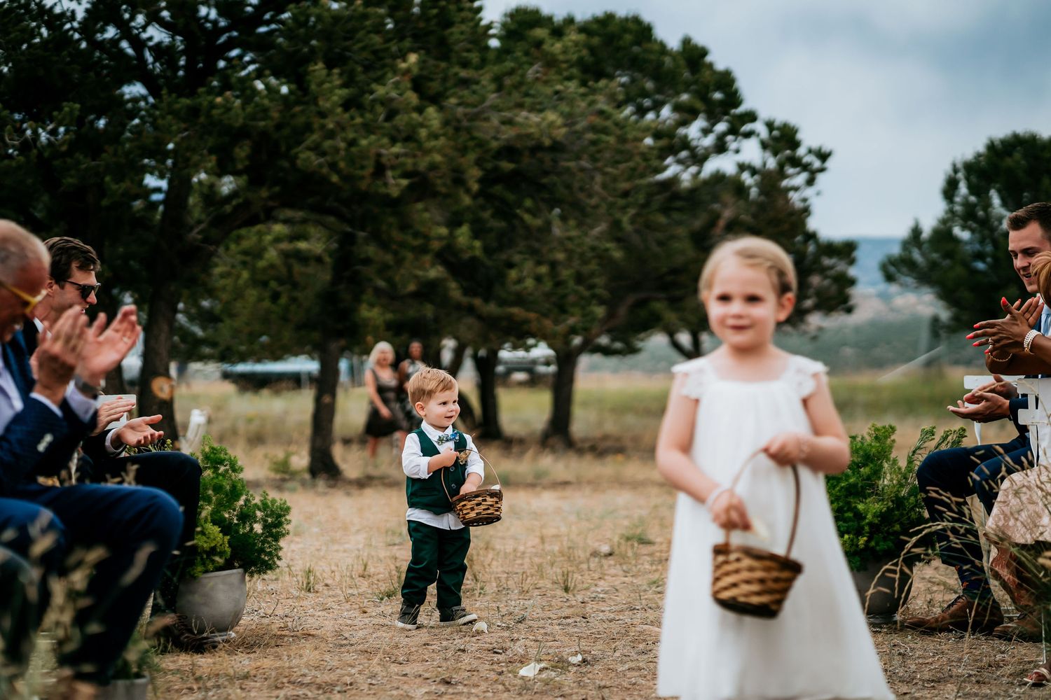 Flower girl in white dress walks down aisle at outdoor mountain wedding ceremony.
