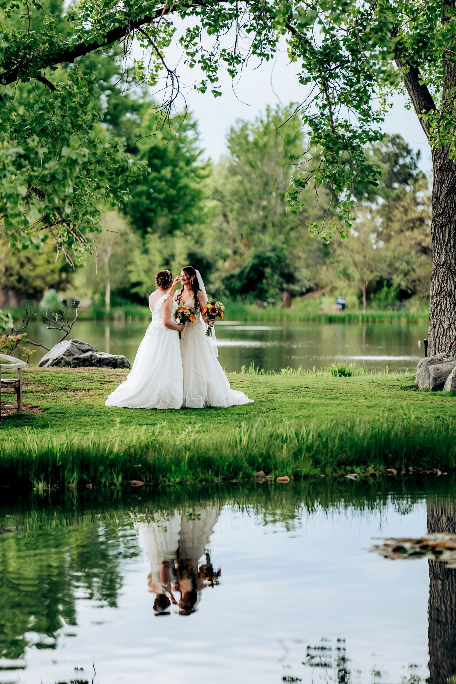 Wedding couple poses by tranquil pond with their reflection visible in still water under green trees.