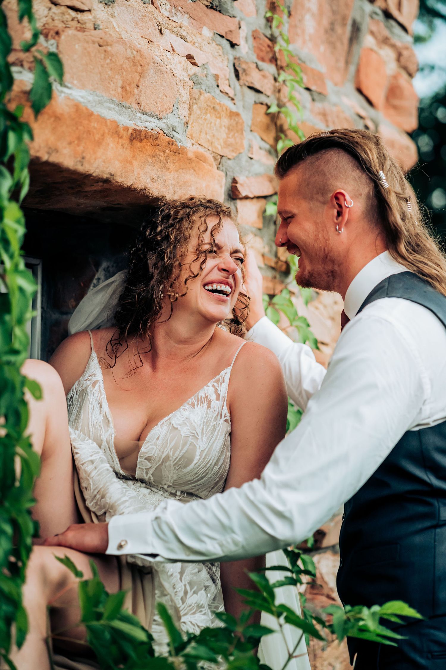 A joyful wedding moment shared between partners against an old brick wall with climbing vines.