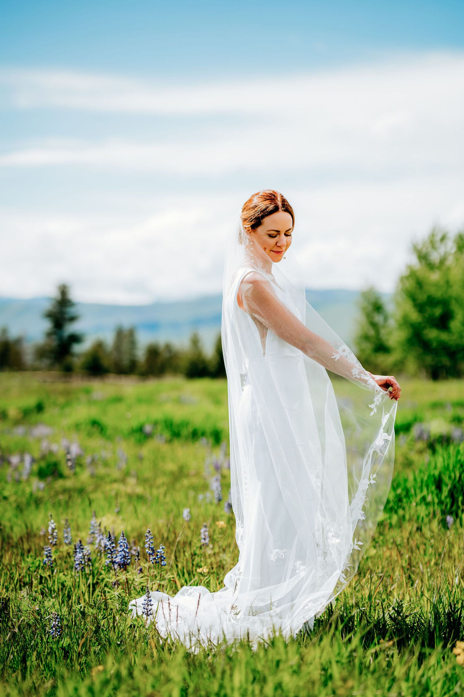 Elegant bride poses with flowing veil in a mountain meadow with wildflowers and blue skies.