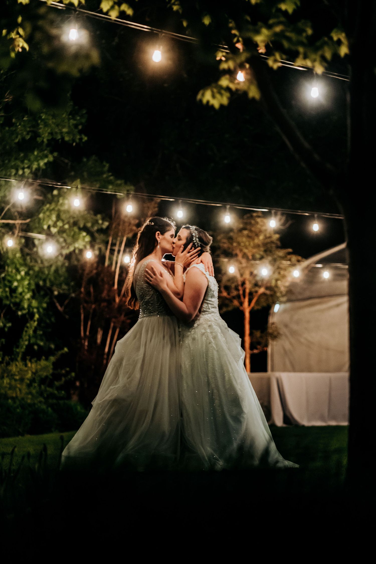A romantic moment under string lights at a nighttime outdoor wedding celebration.