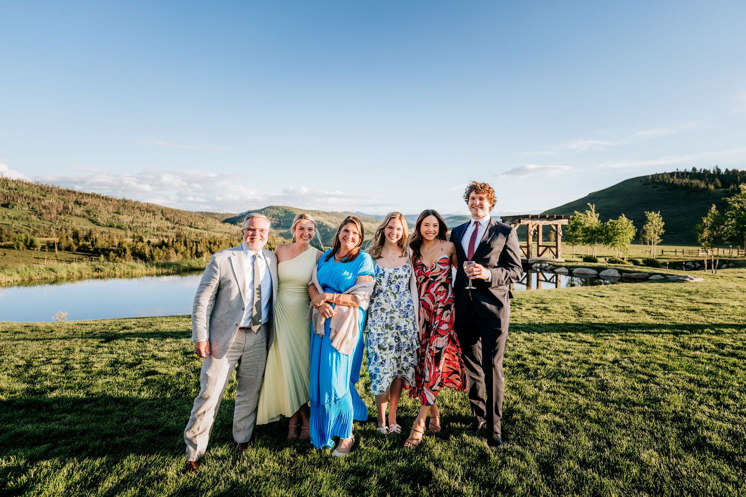 Group poses together on grassy hill with scenic lake and mountains in background at outdoor event.
