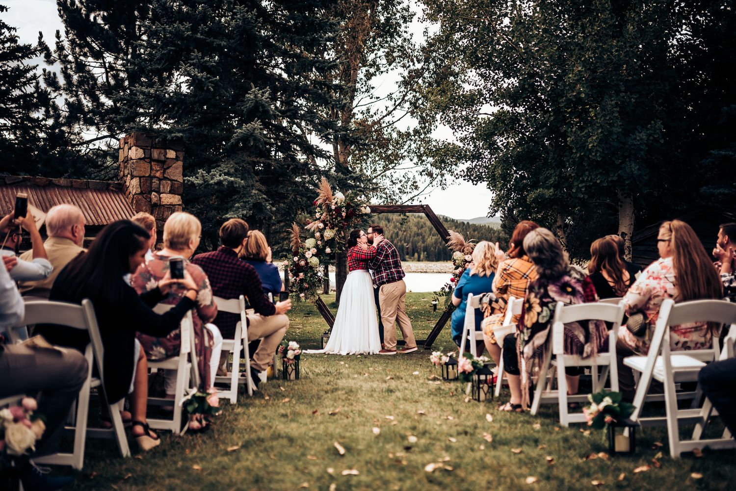 Outdoor wedding ceremony setup with white chairs and guests seated on a grassy lawn surrounded by trees.