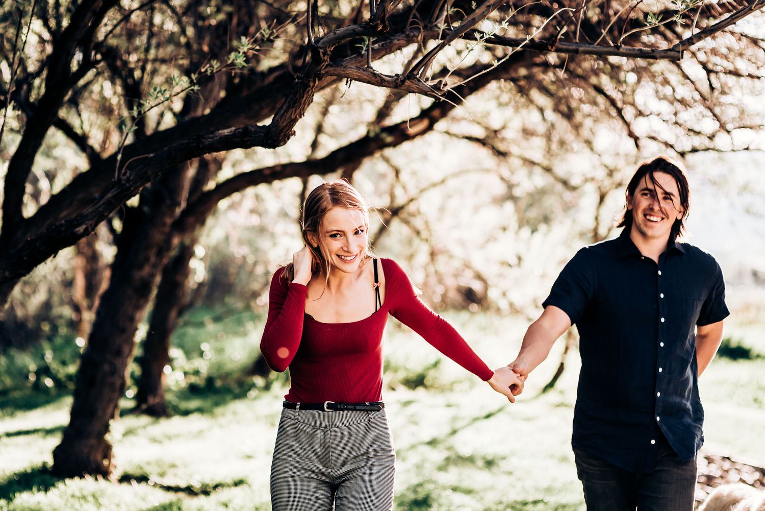 Couple holds hands while walking through sunlit forest path.