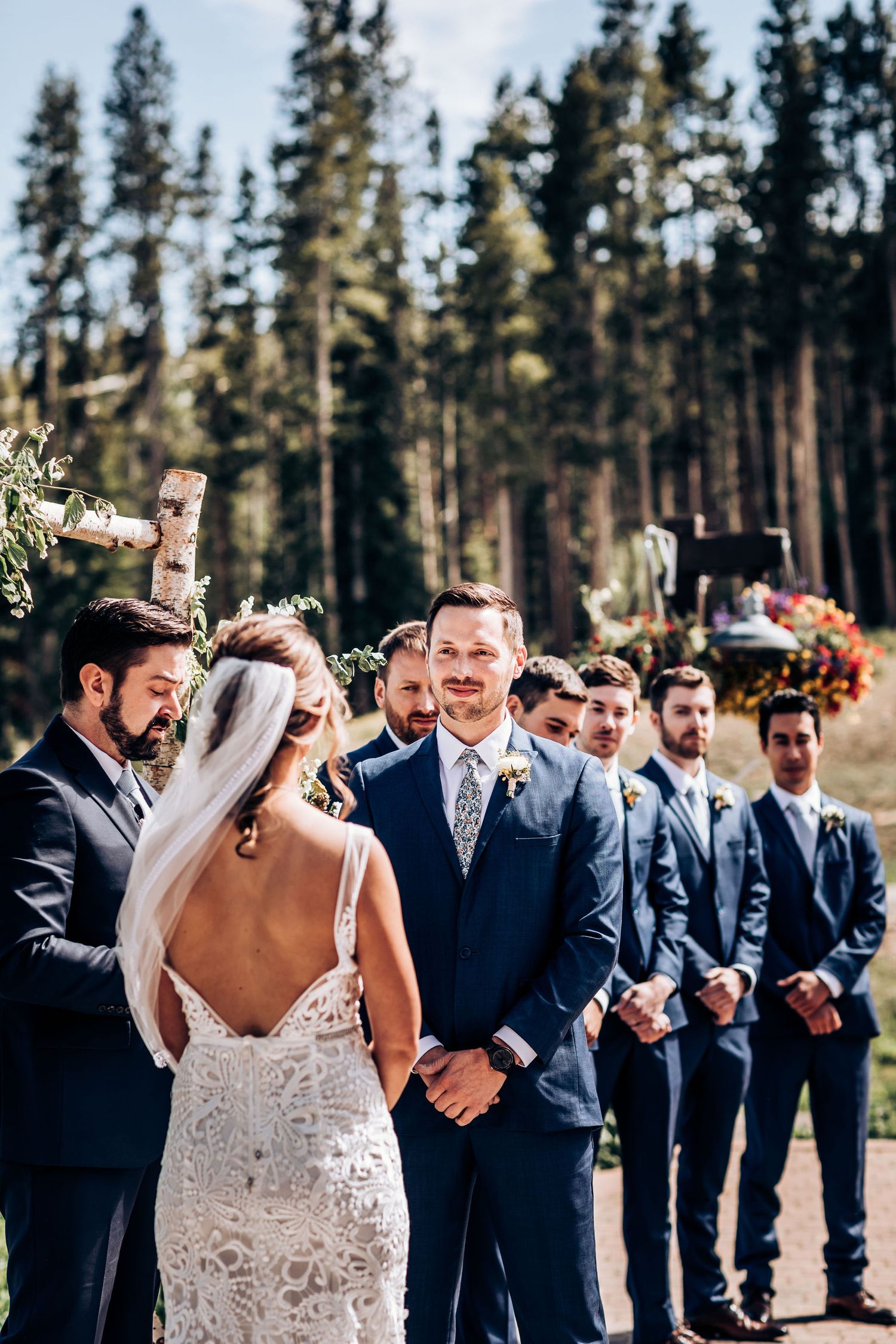 Wedding party stands in formation during outdoor ceremony with tall pine trees in background.