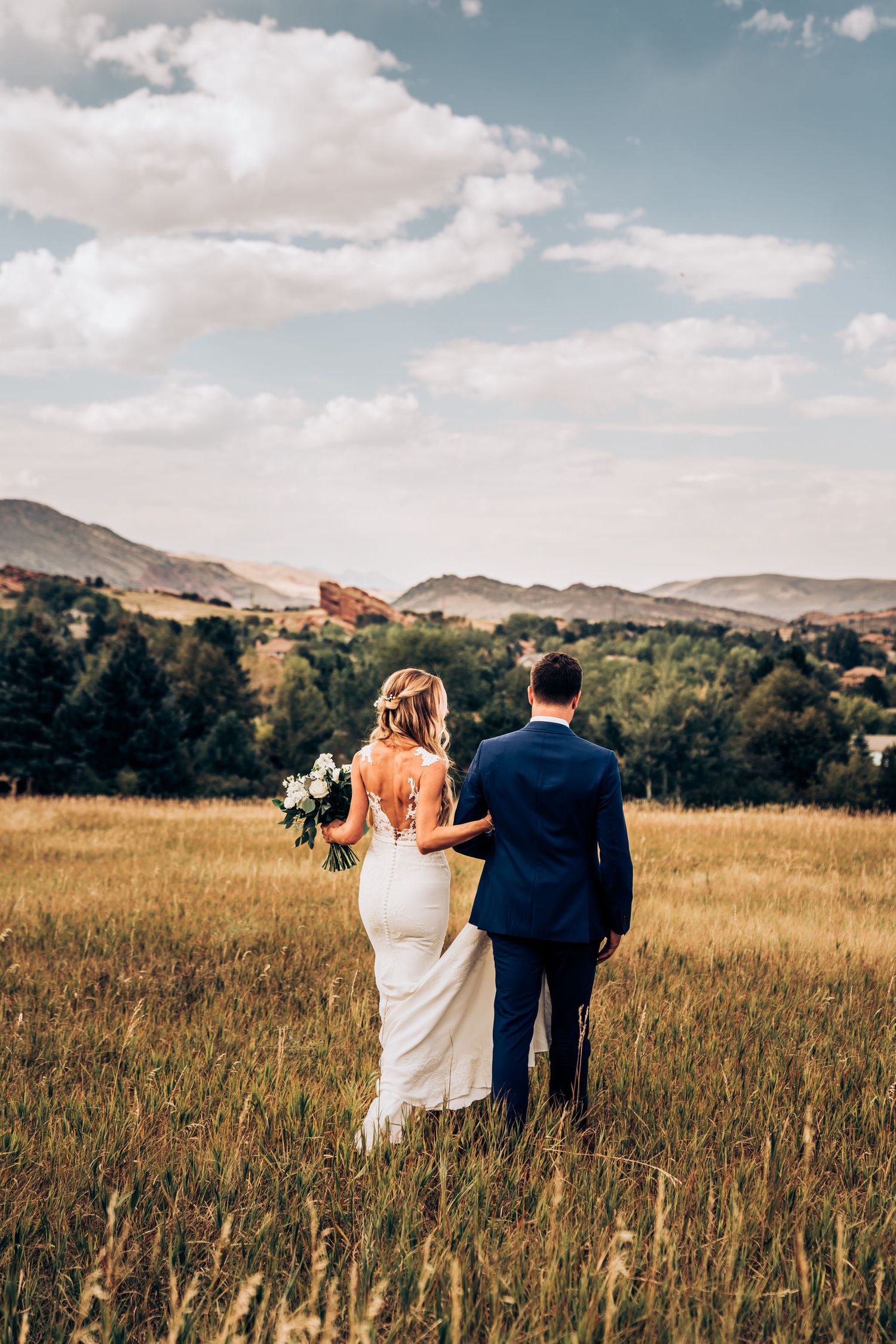 Couple walks through golden mountain meadow with white bouquet and formal attire.