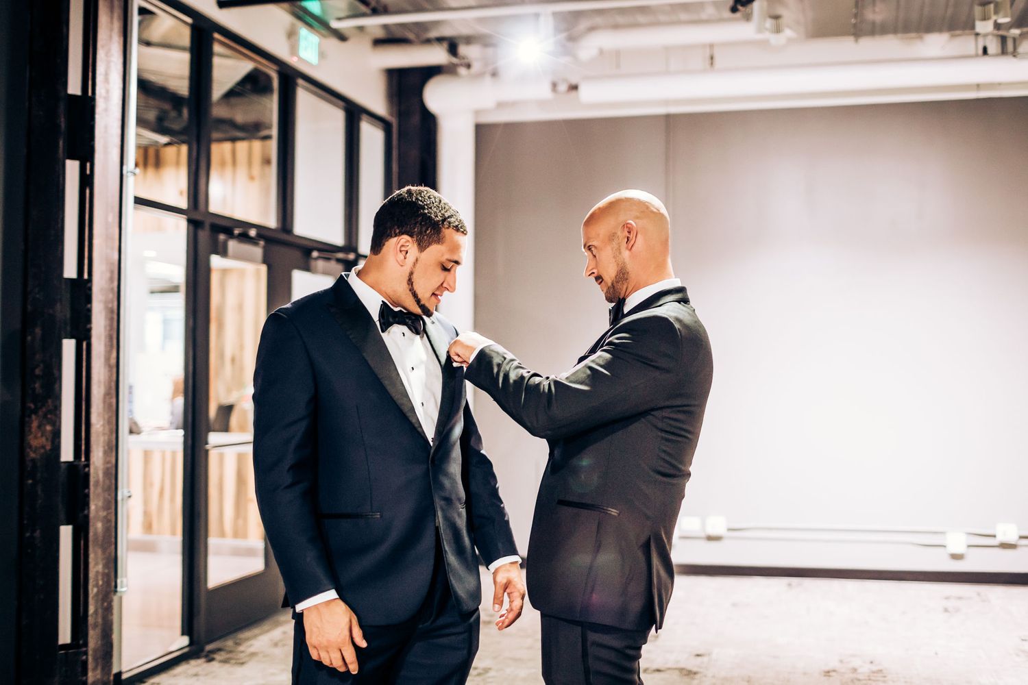 Two groomsmen in black tuxedos adjust formal attire before ceremony.