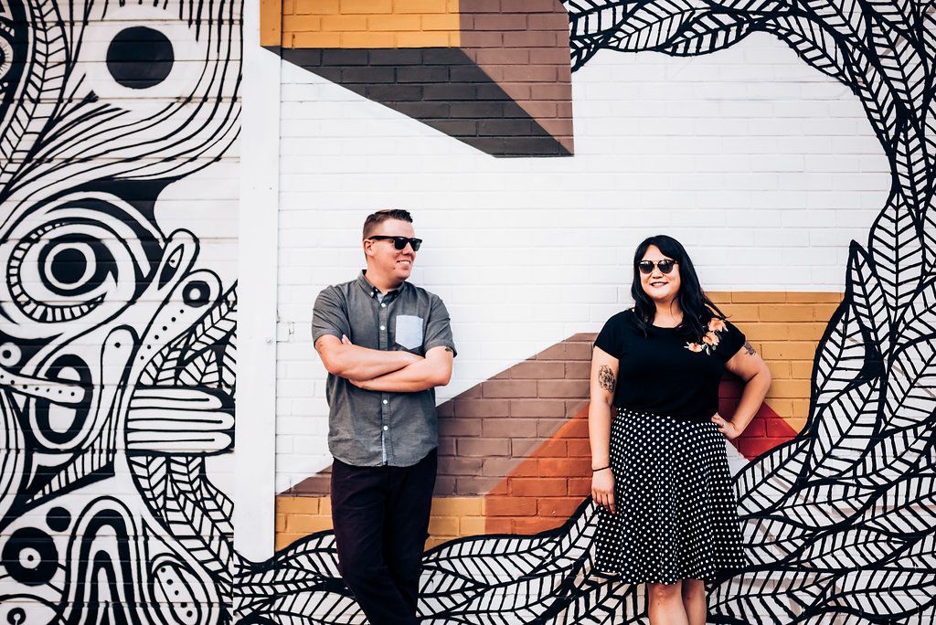 A stylish pair posing against a black and white geometric mural on a brick wall.