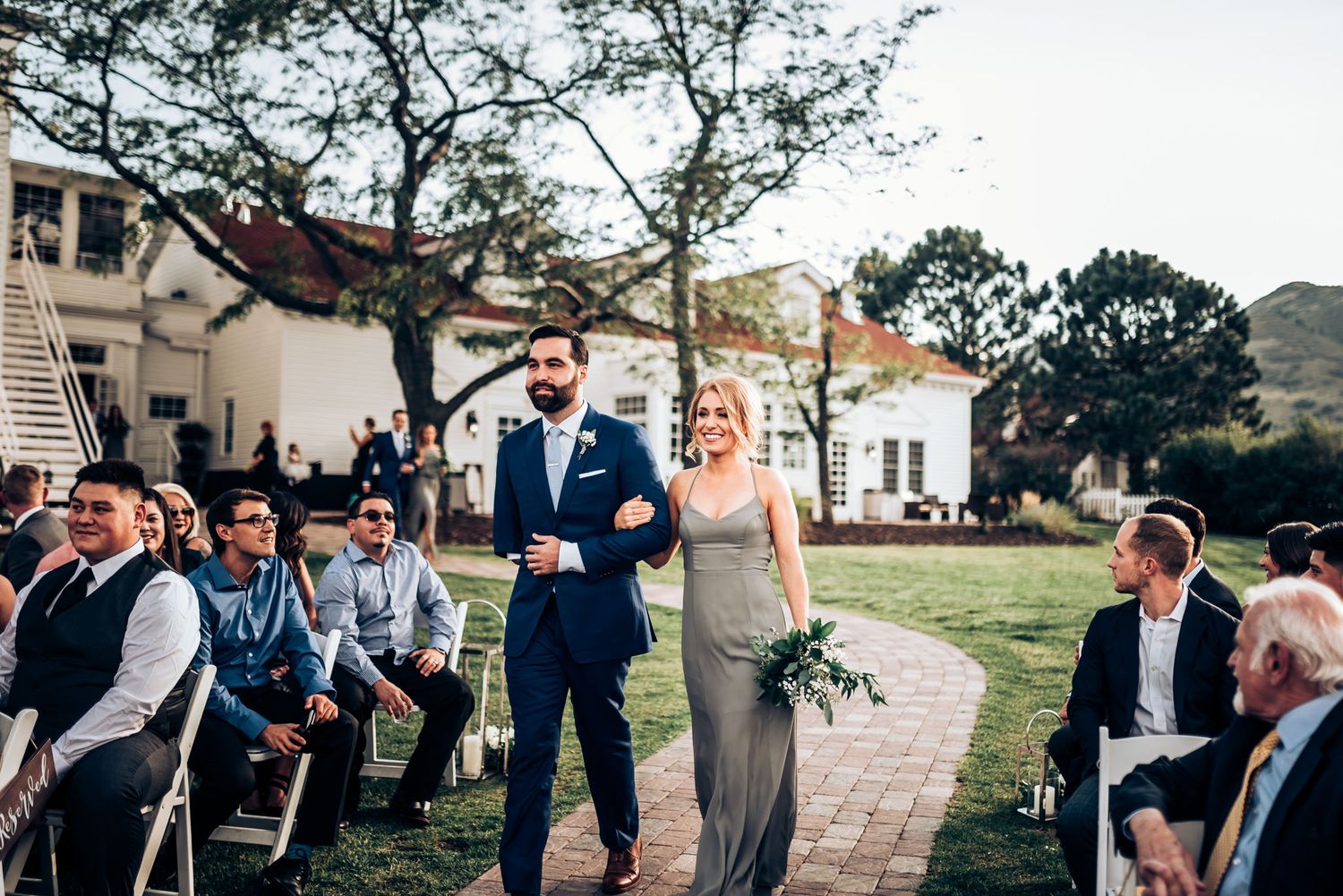 Couple walking down stone aisle at outdoor wedding ceremony with mountain backdrop.