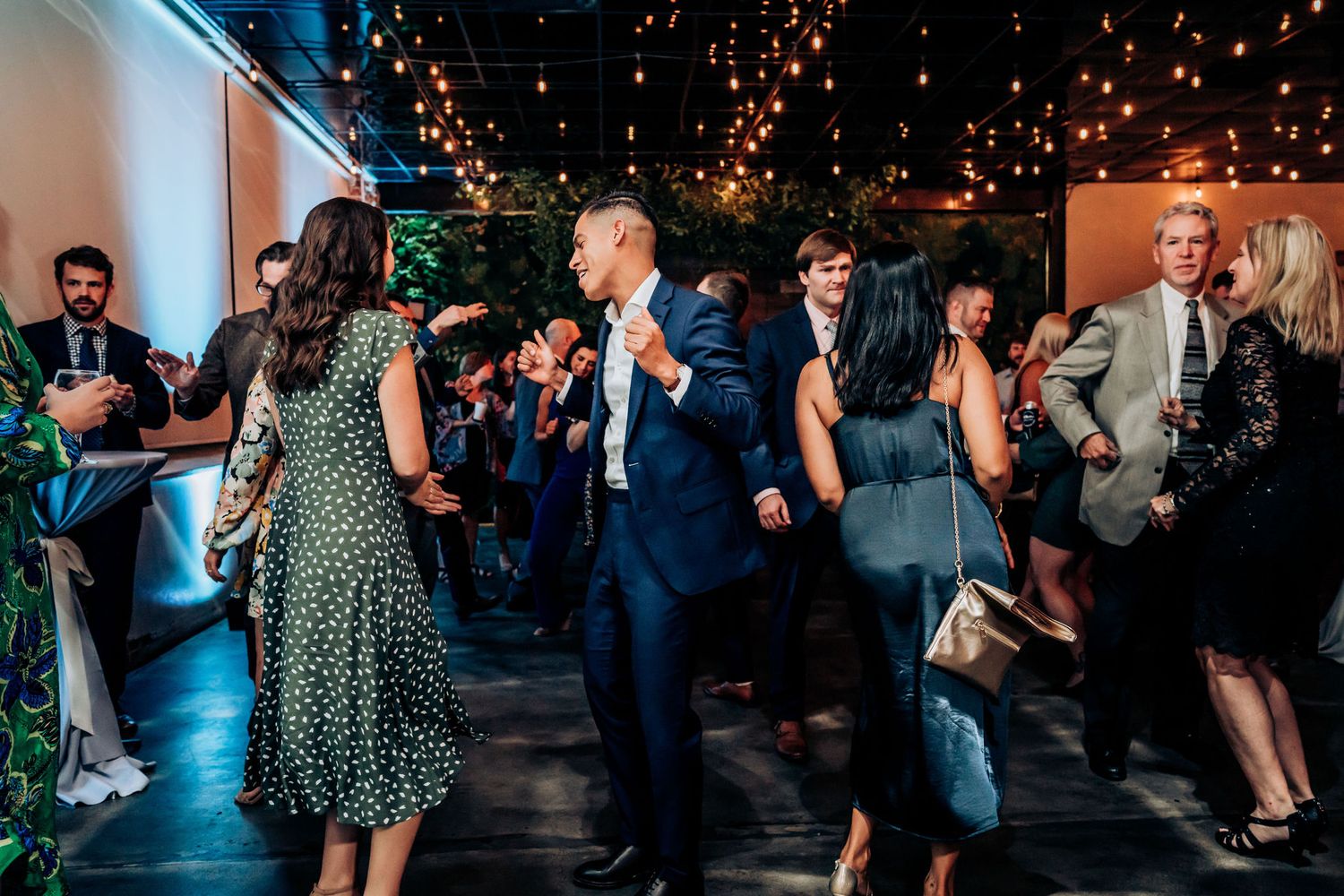 Wedding guests dancing and celebrating under twinkling string lights at an indoor reception venue.