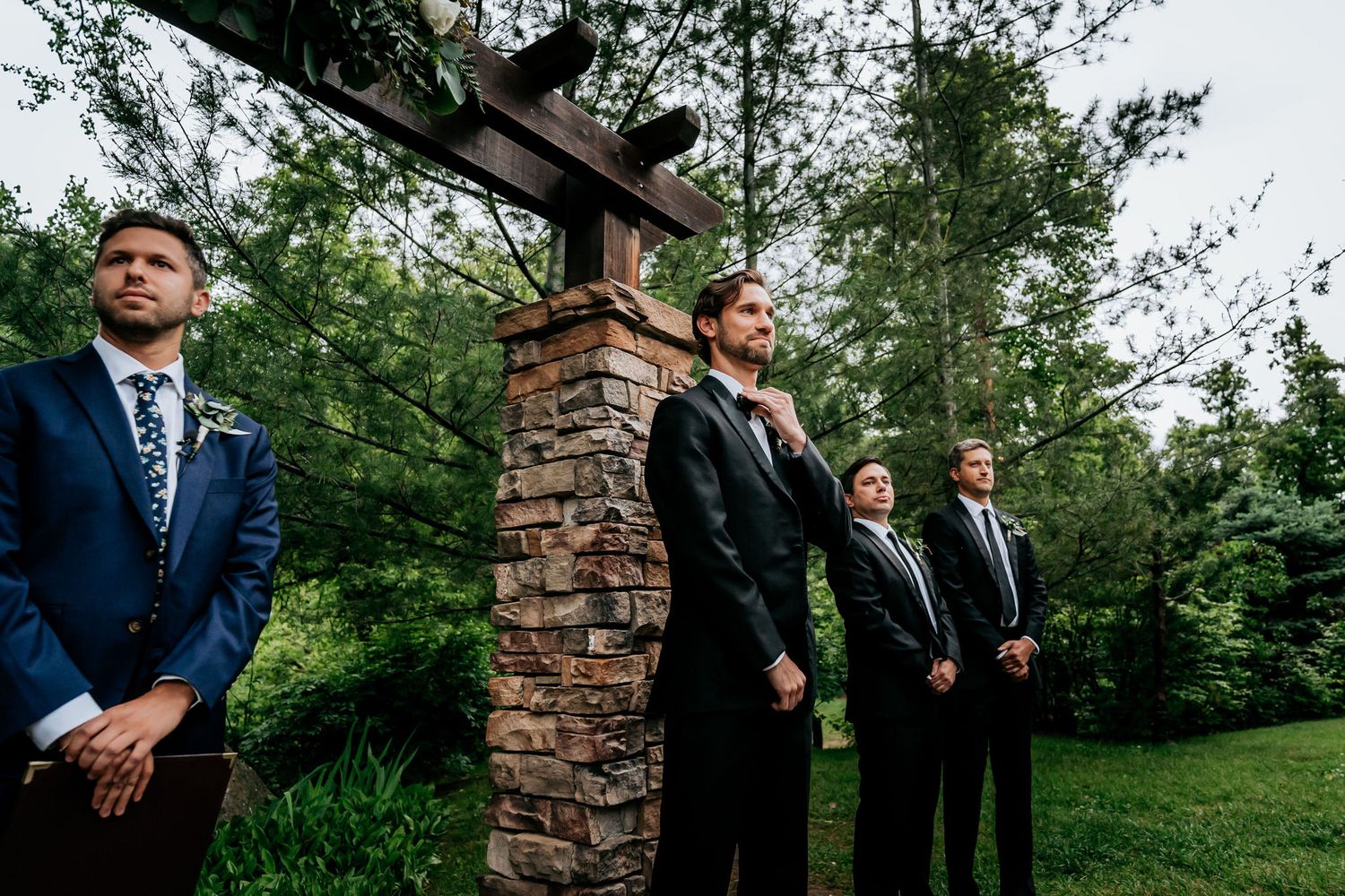 Wedding party members stand in formation by a stone pillar during an outdoor ceremony.