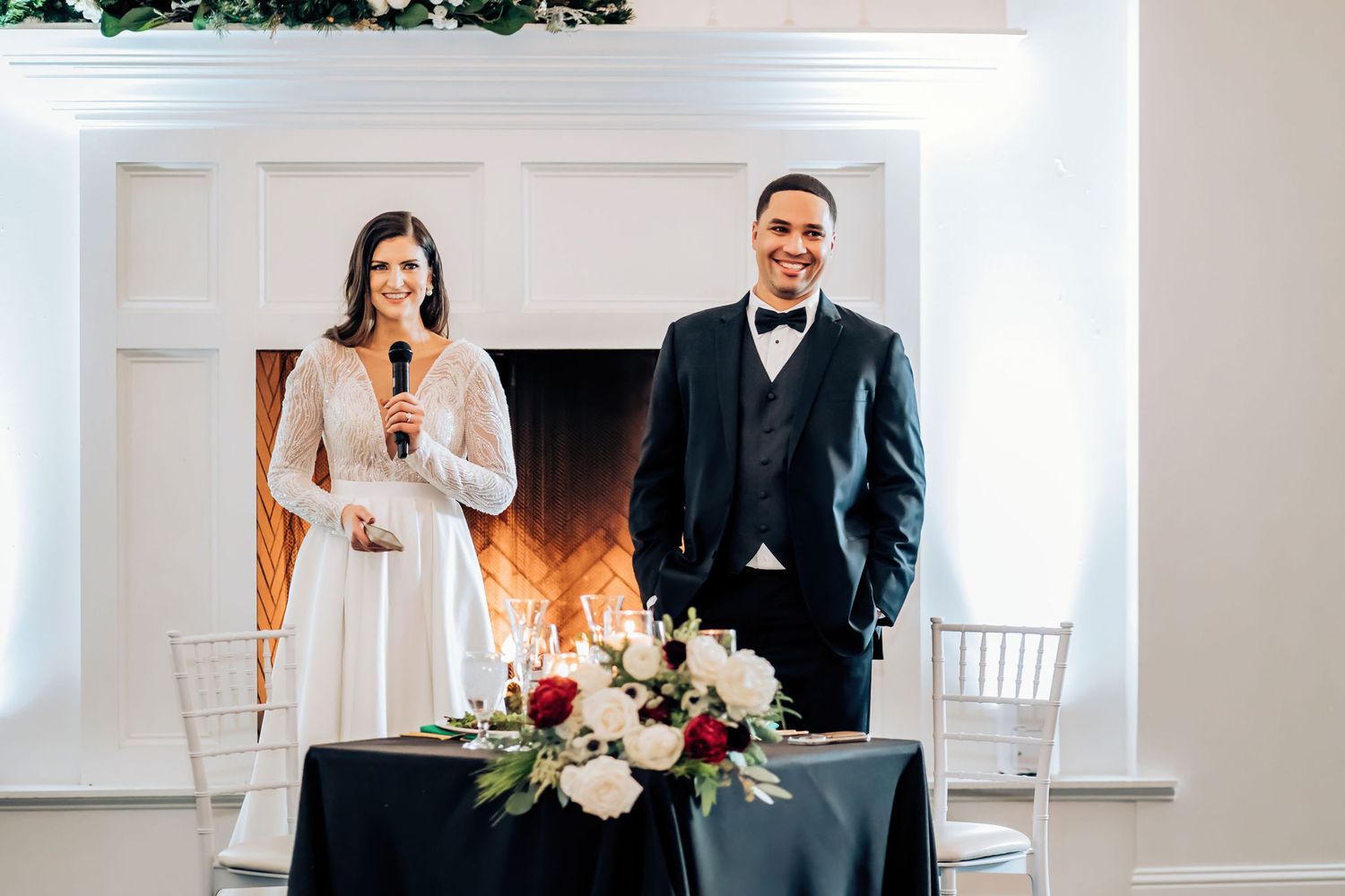 Newlyweds stand at decorated head table with fireplace and floral arrangements.