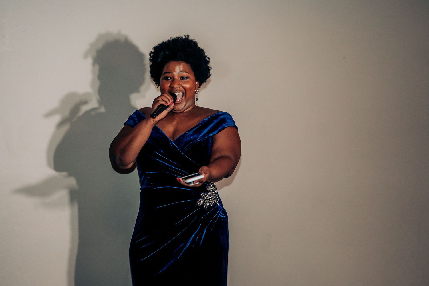 Wedding performer in a blue velvet dress sings against a white wall with dramatic shadow.