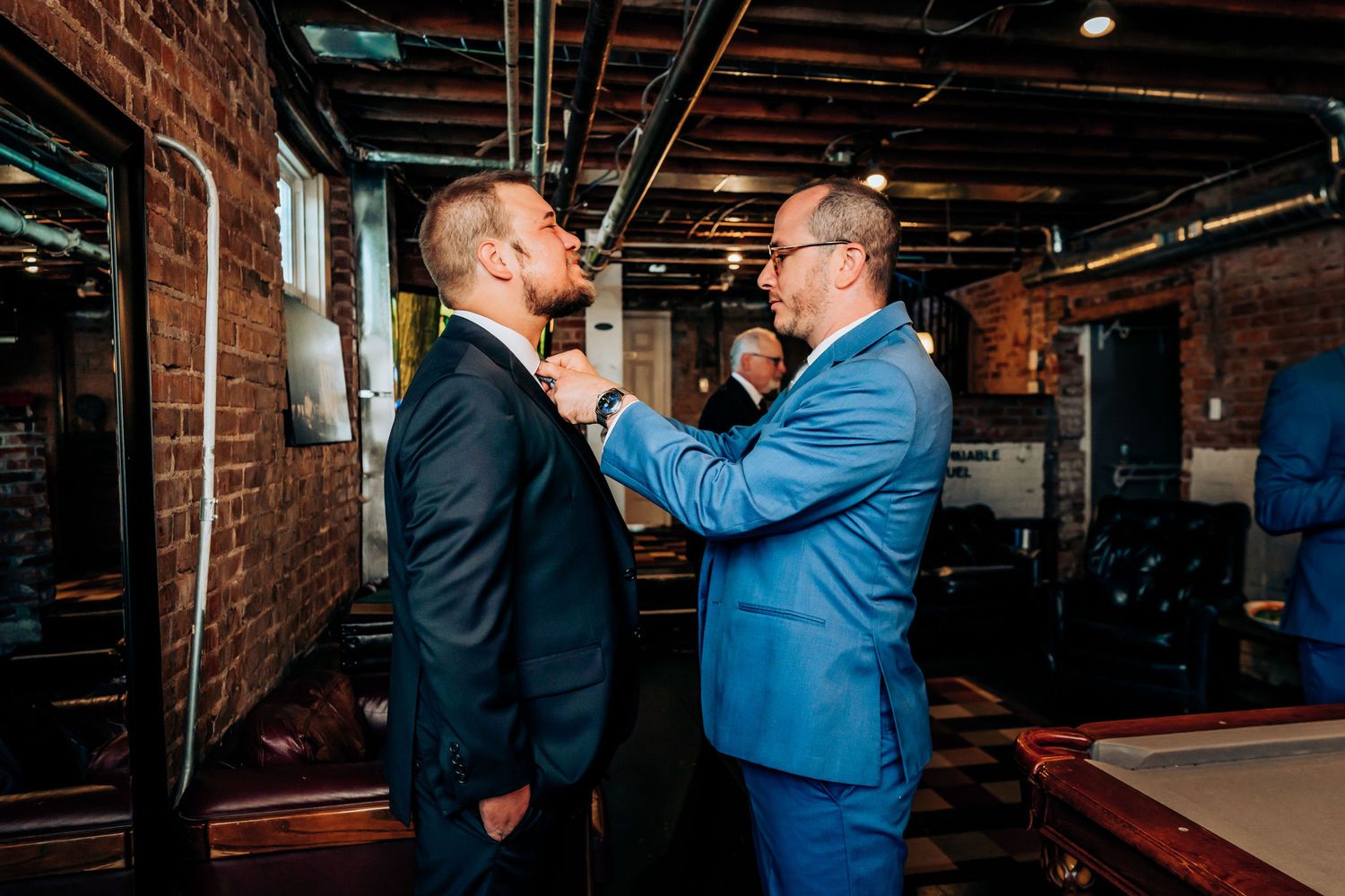 Person helps adjust another's tie in industrial-style room with brick walls.