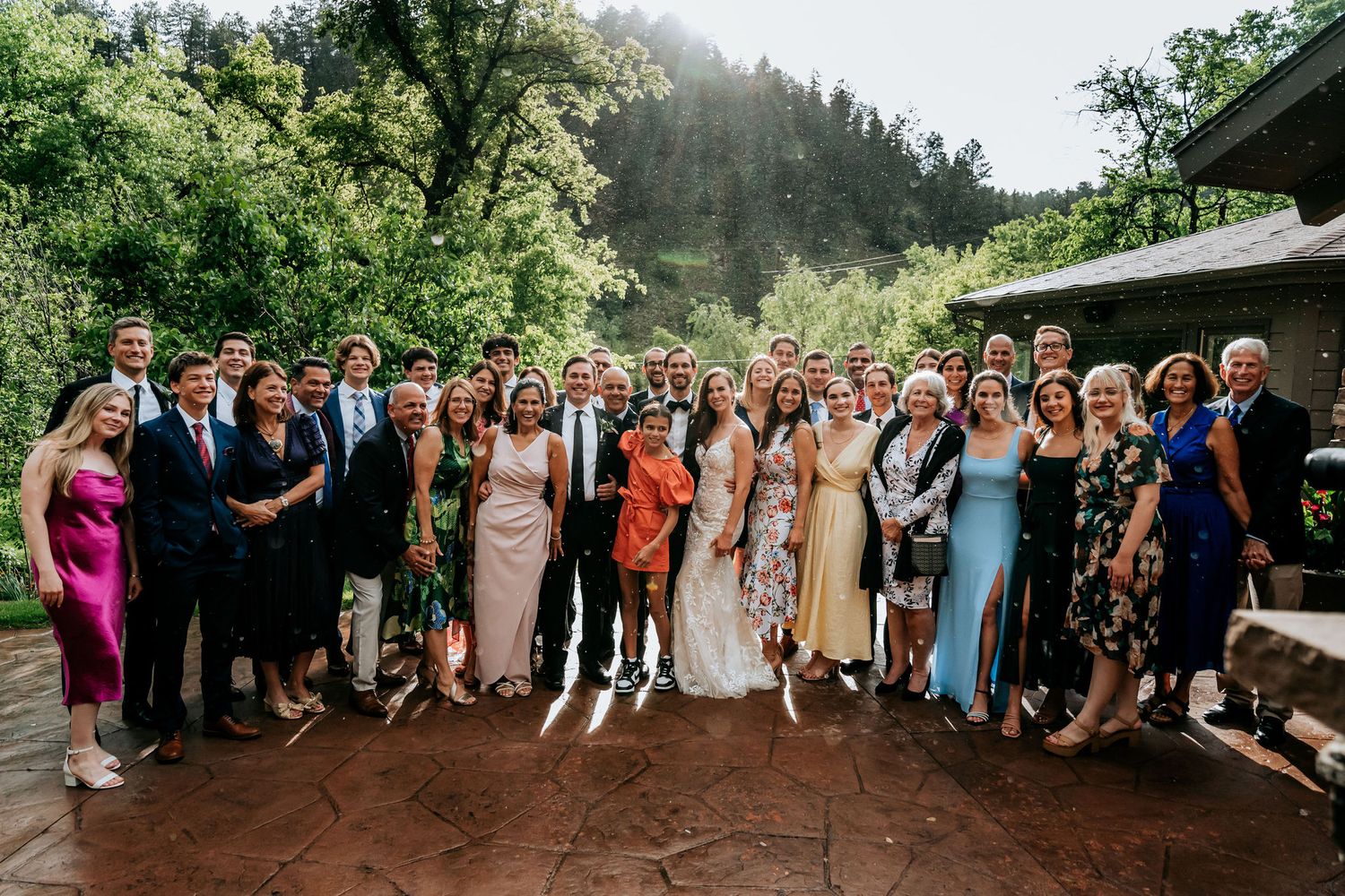 Large group photo of wedding guests on an outdoor terrace surrounded by mountain scenery.
