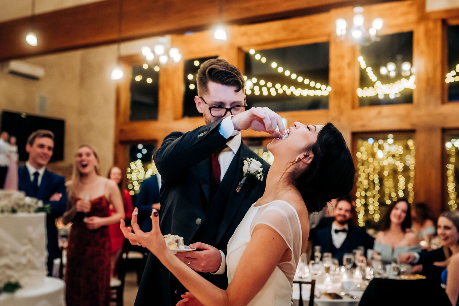A playful cake-feeding moment between newlyweds at their reception with string lights in background.