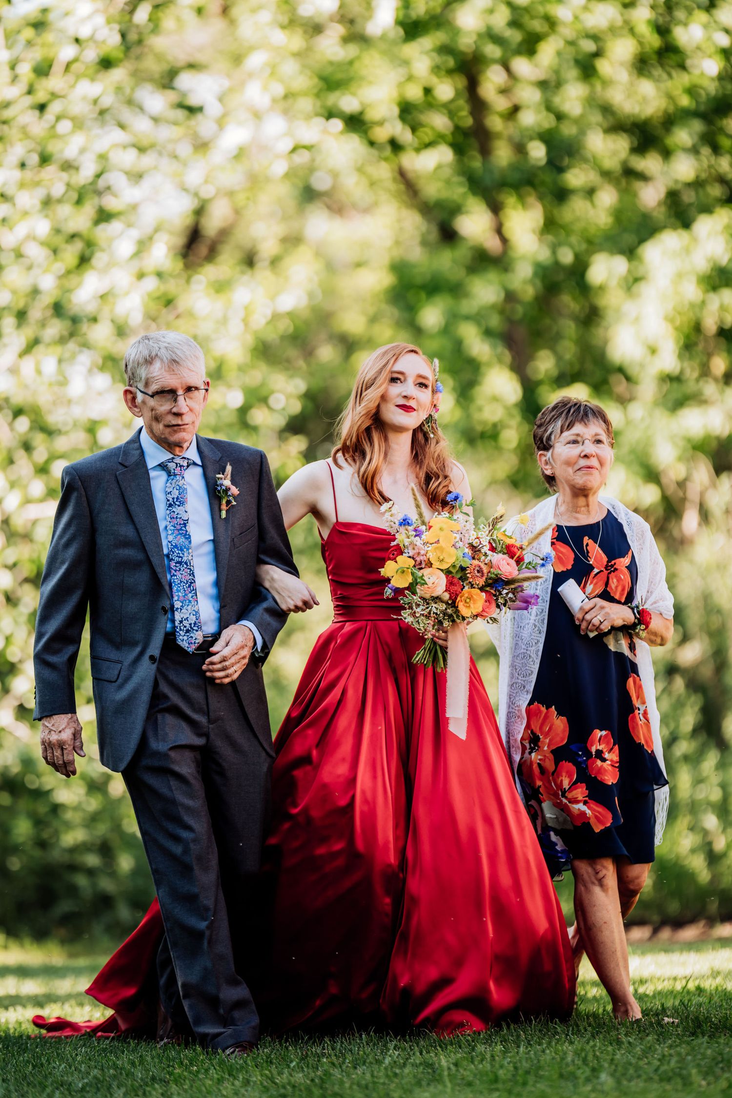 A wedding party walks down a garden path, featuring a stunning red ball gown and formal attire.