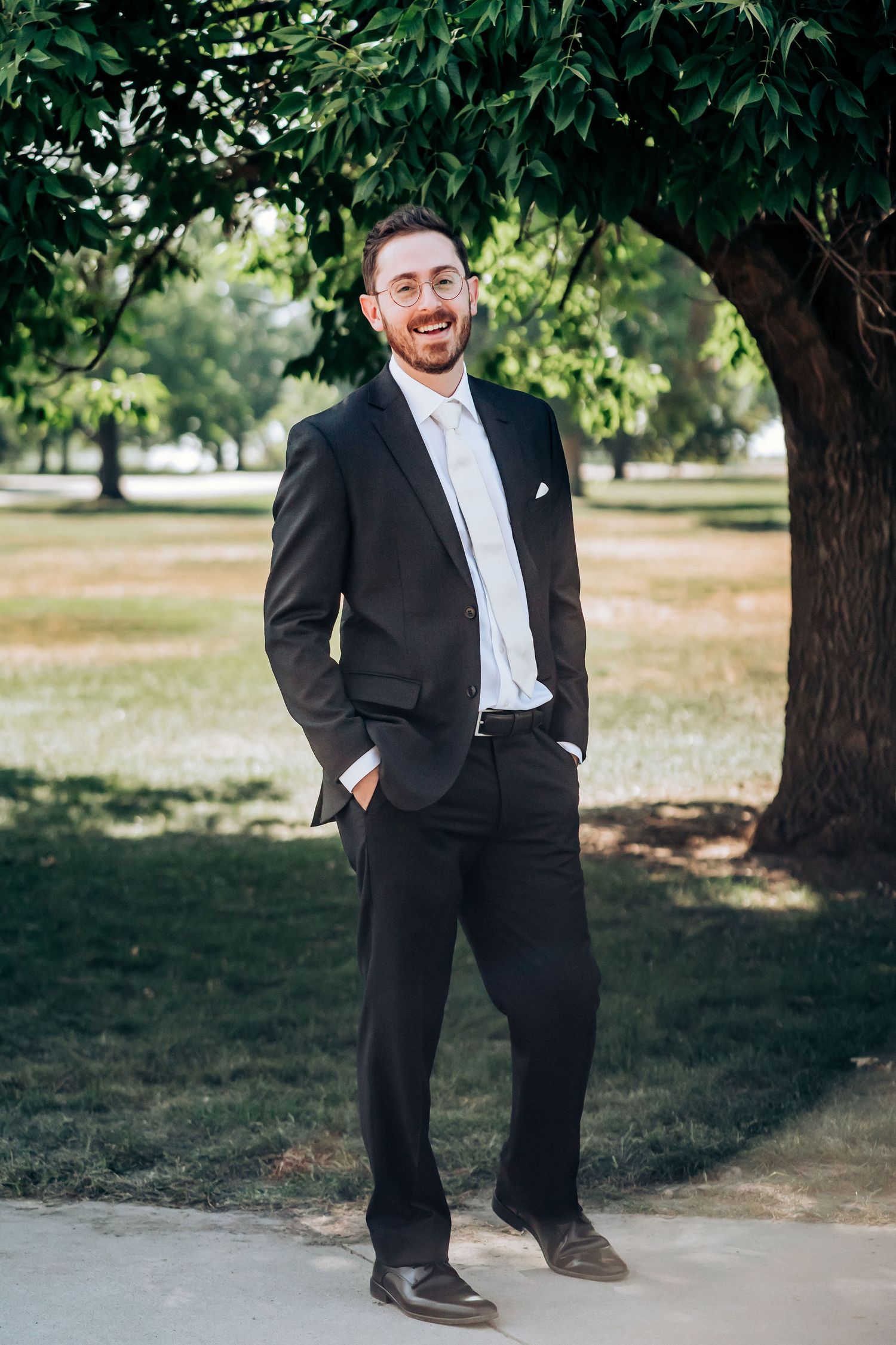 A person in a classic black suit poses casually under trees in an outdoor wedding setting.