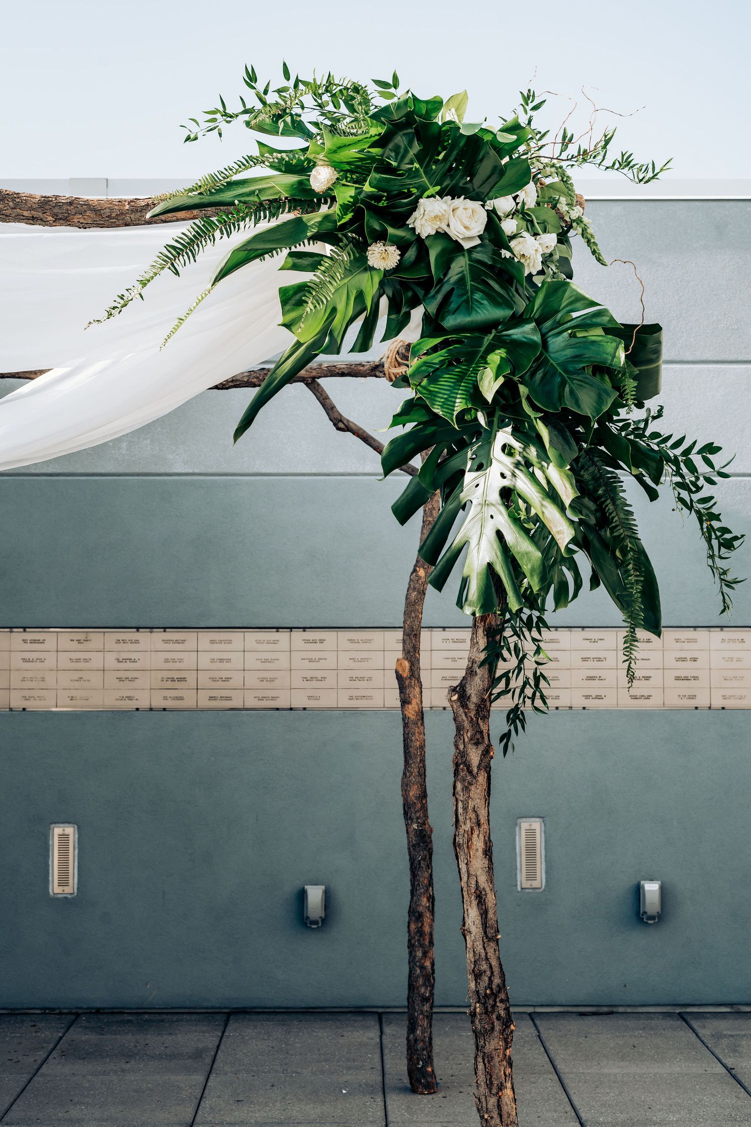Modern wedding arch decorated with tropical leaves and white flowers against industrial wall.