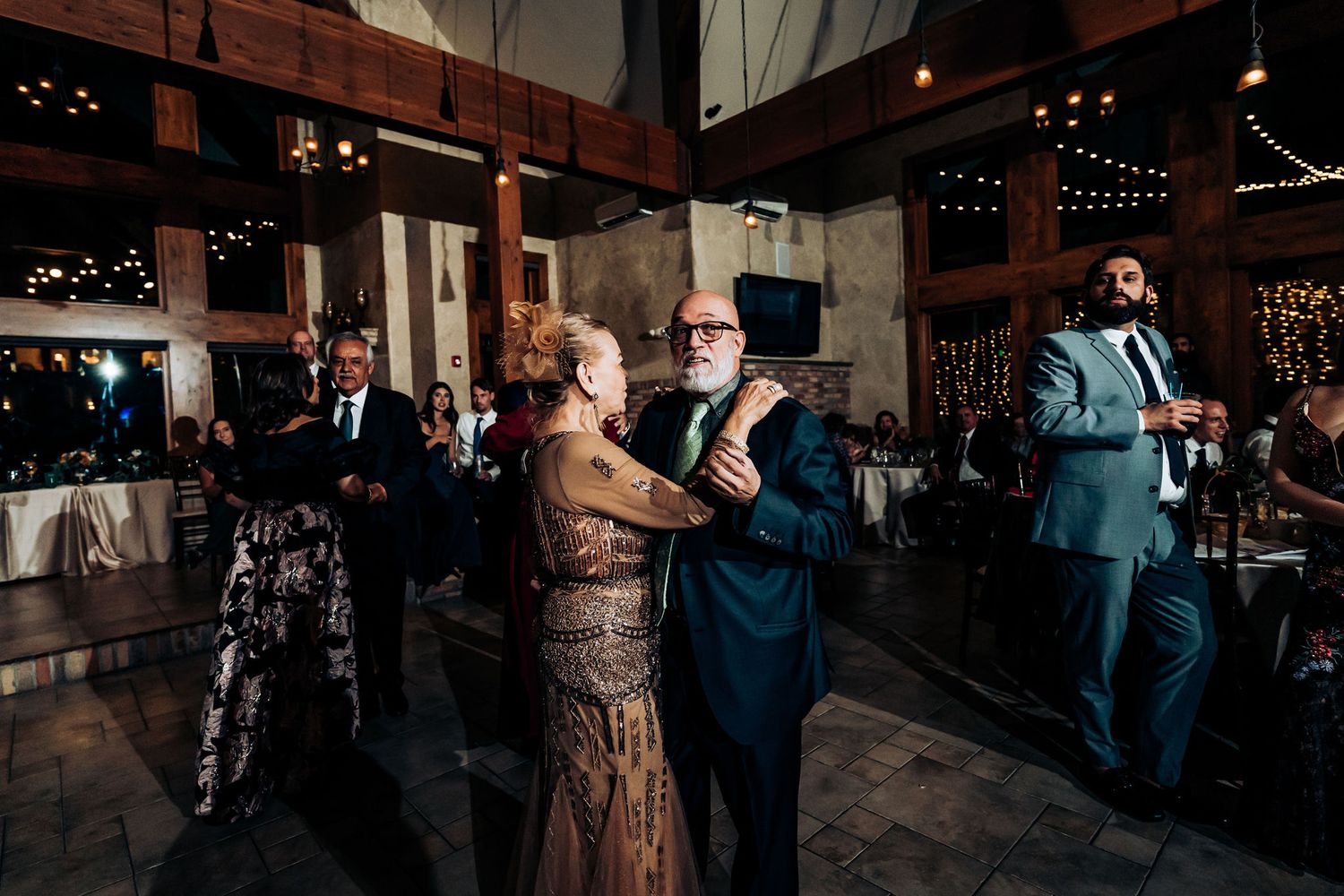 Wedding guests dancing together on wooden dance floor during evening celebration with string lights overhead.