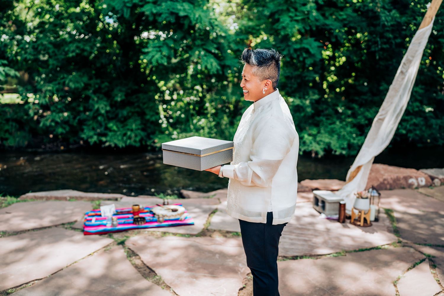 In an outdoor setting with greenery, a person in a cream barong holds a gift box during a ceremony.