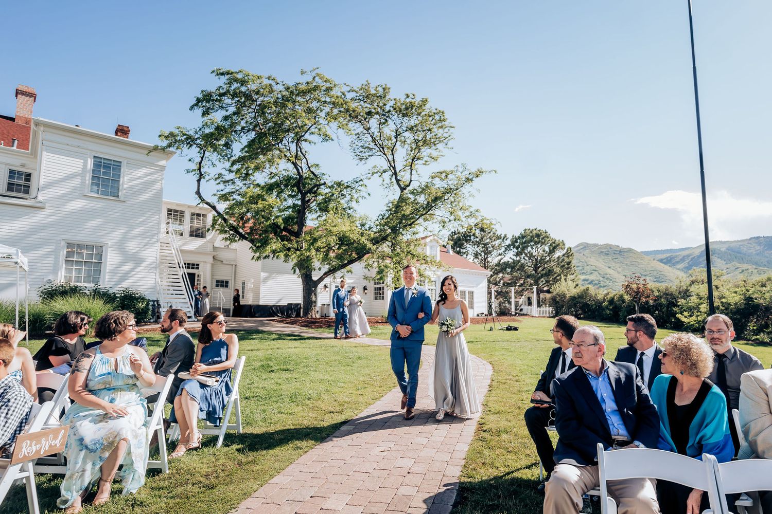 Outdoor wedding ceremony with guests seated on lawn in front of white mansion.