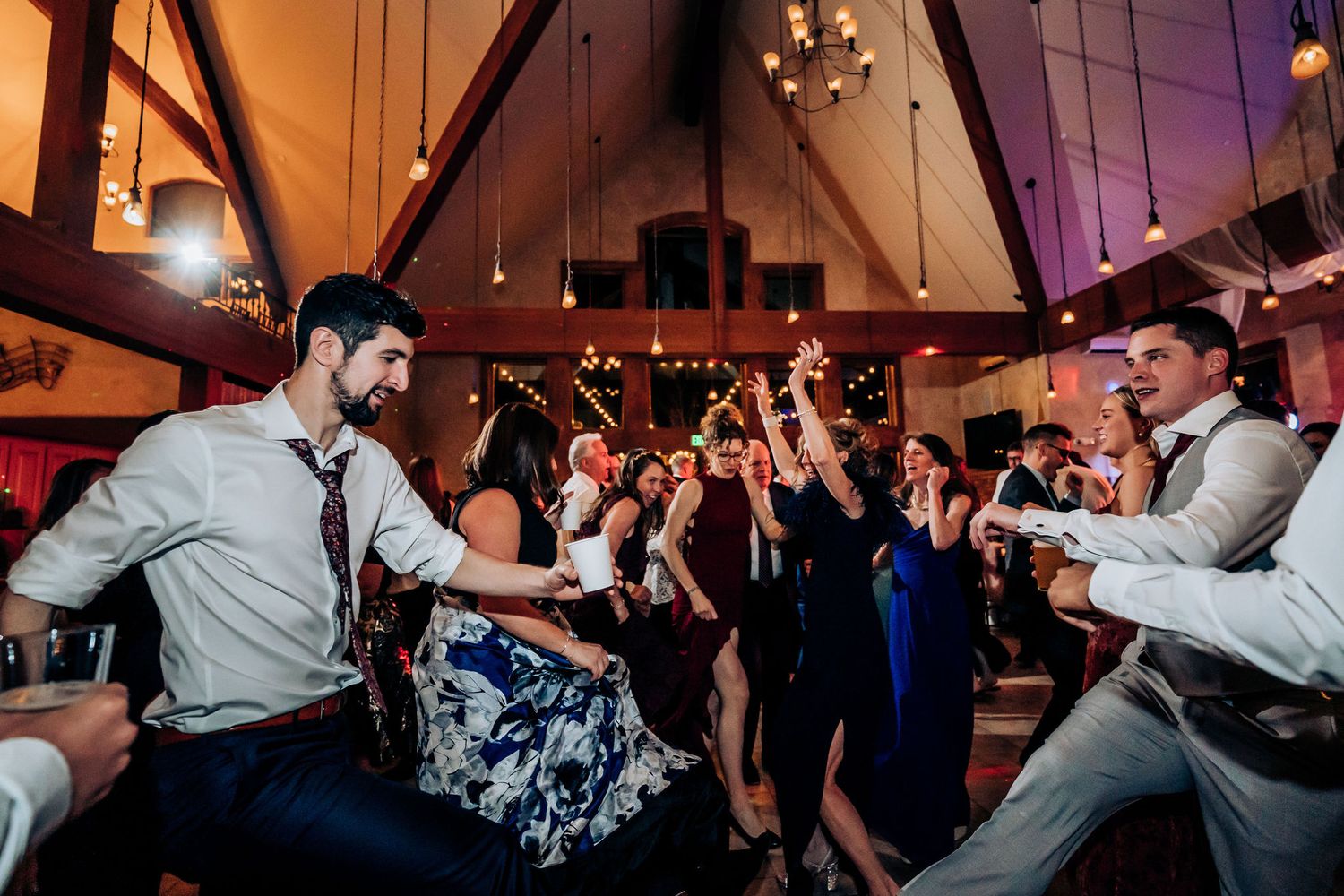 Wedding guests dance energetically at a rustic venue with vaulted ceilings and warm lighting.