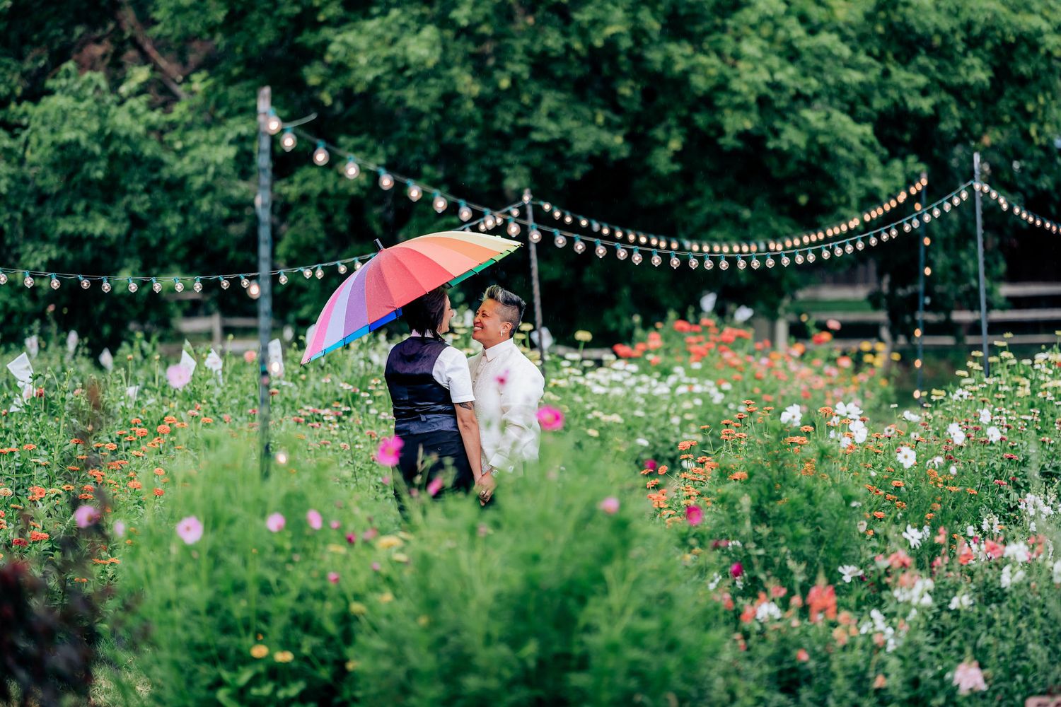 A couple shares a rainbow umbrella in a wildflower garden with string lights above.