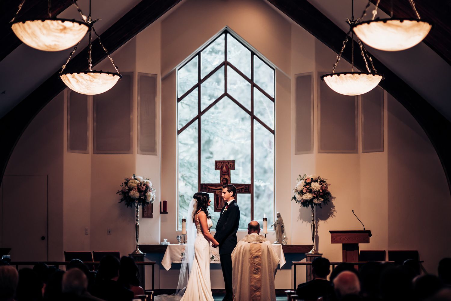 Wedding ceremony in a modern church with vaulted ceilings, ornate lighting fixtures, and a large geometric window.