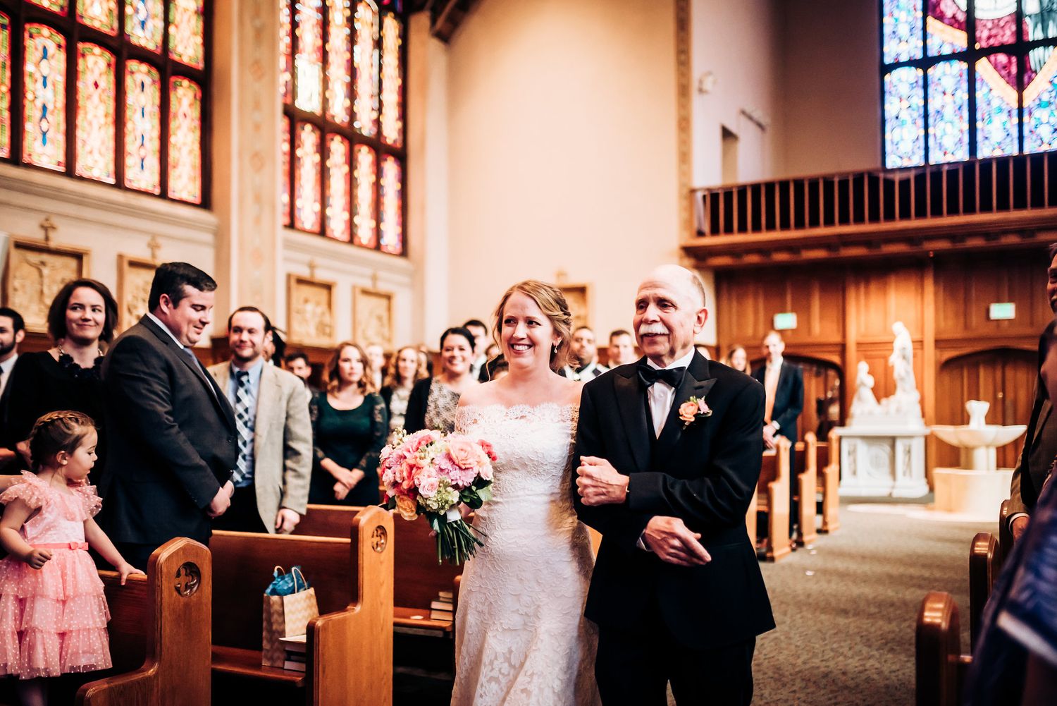A wedding procession walks down the aisle of a traditional church with stained glass windows.