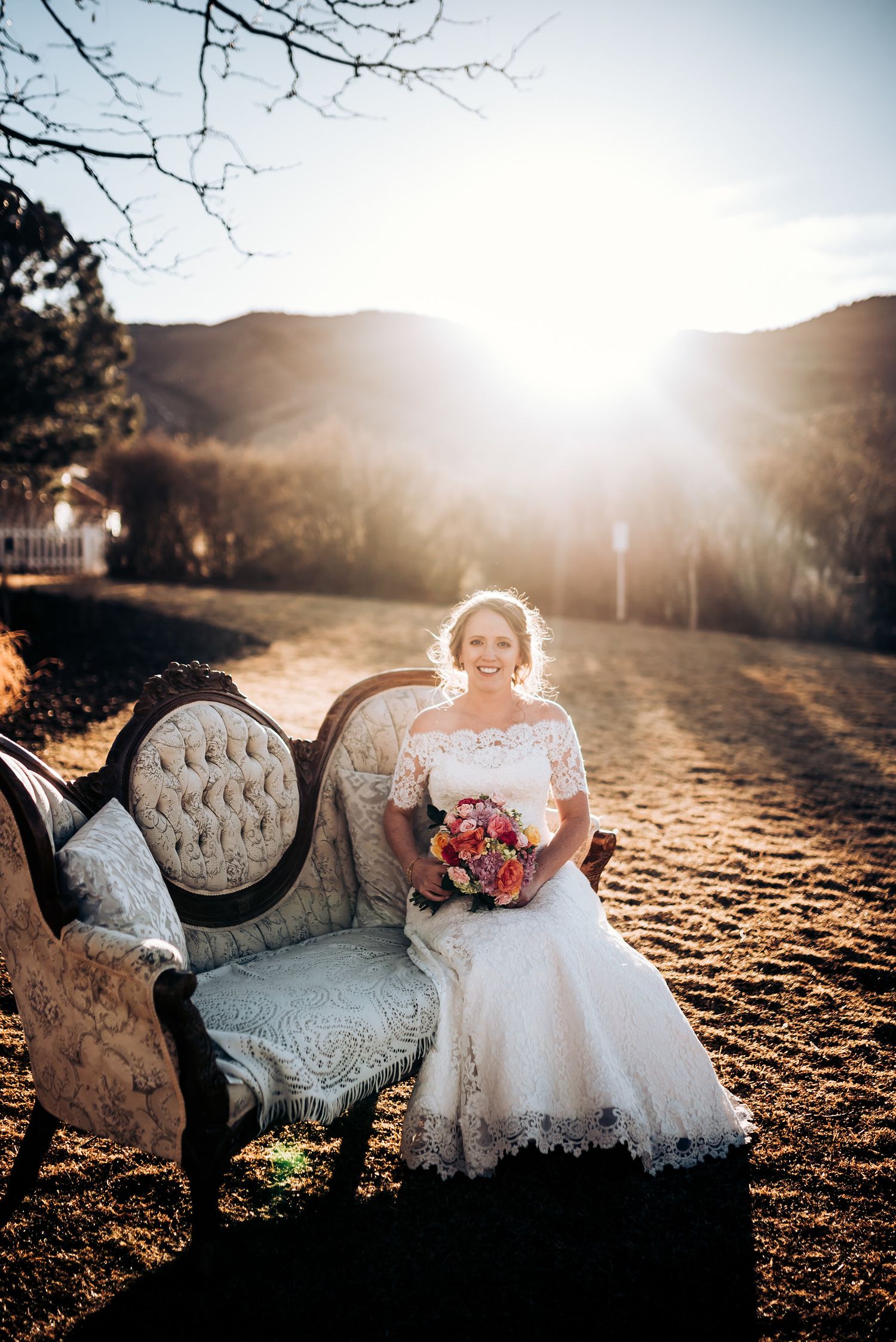 Bride seated on ornate vintage couch outdoors holding vibrant bouquet with mountain backdrop.