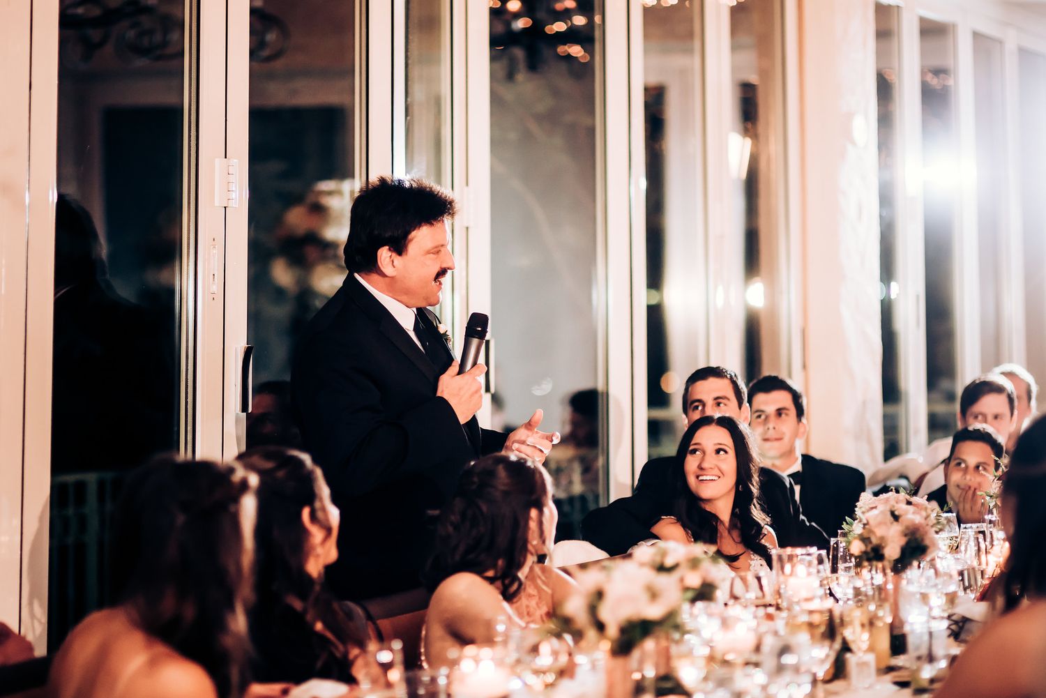 Speaker stands to deliver wedding toast while guests listen attentively at decorated reception tables.
