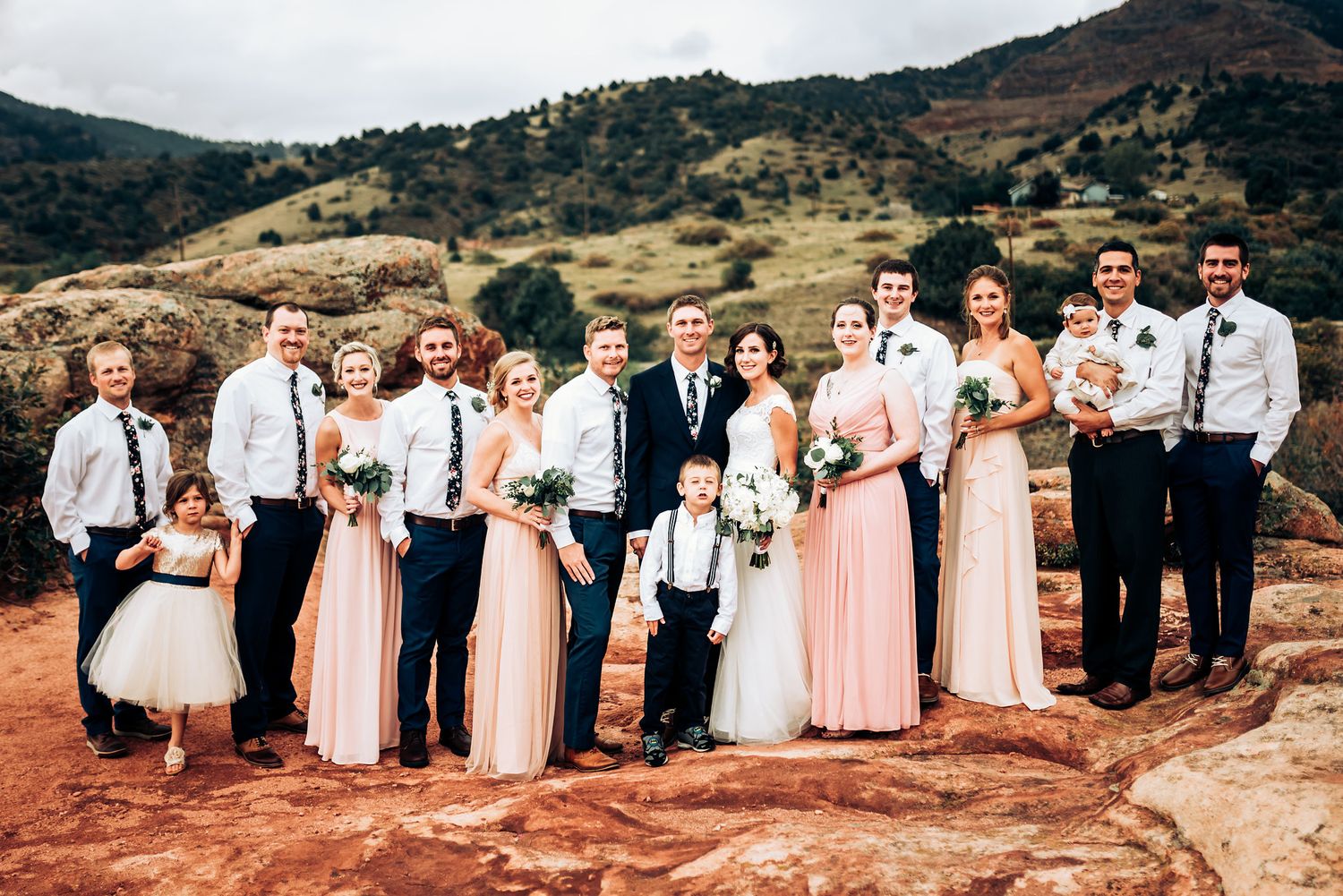 Large wedding party in formal attire gathered on red rock formation with mountain views.