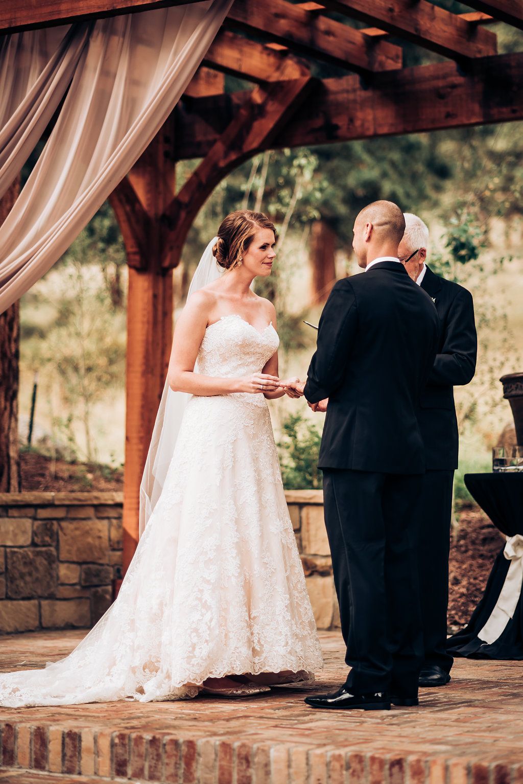 Elegant outdoor wedding ceremony beneath wooden pergola with flowing fabric and stone details.