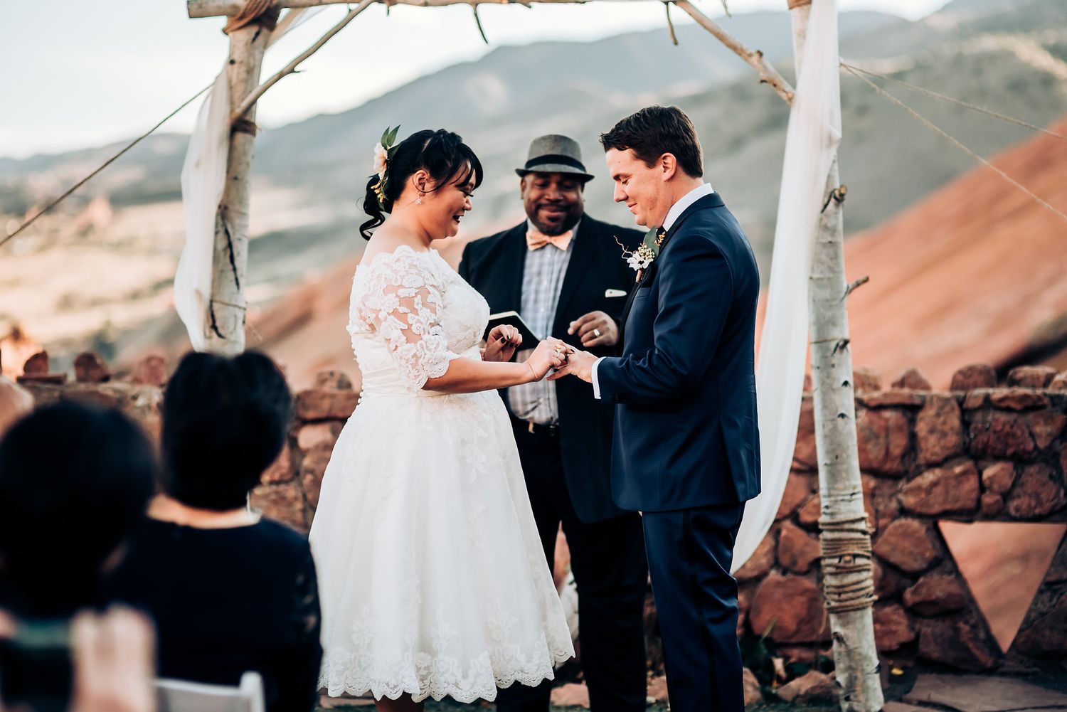 A wedding ceremony takes place under a rustic birch arch with red rock cliffs in the background.
