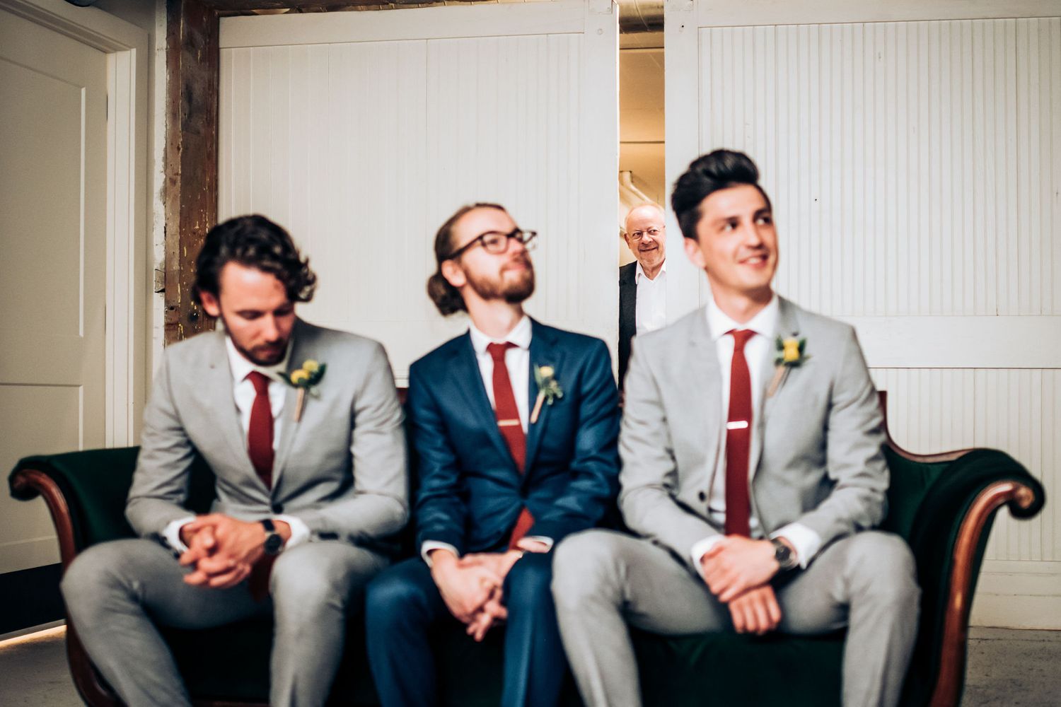 Three groomsmen in matching grey suits and red ties posing casually on couch.