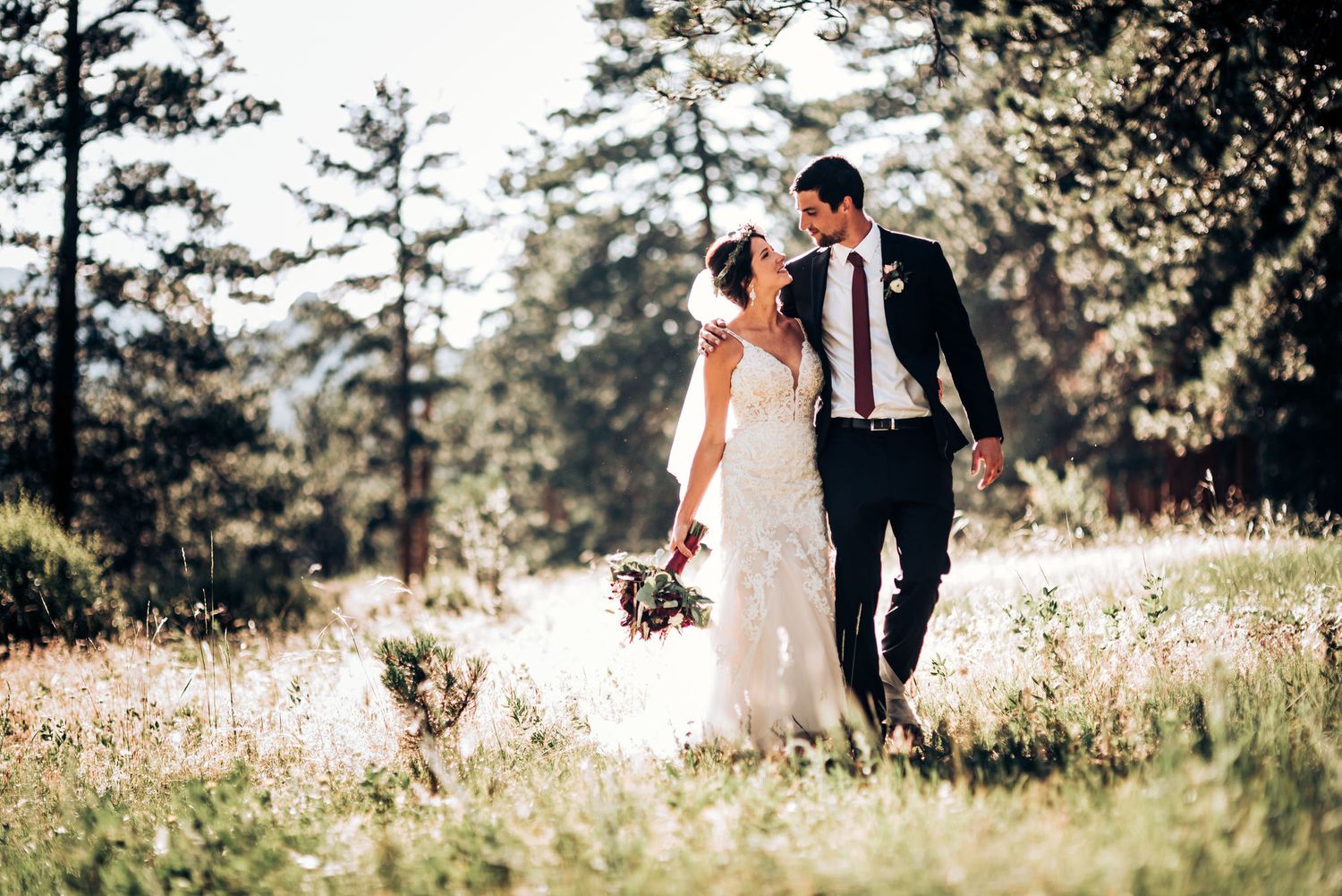 Newlyweds walk hand in hand through a sunny mountain field surrounded by pine trees.