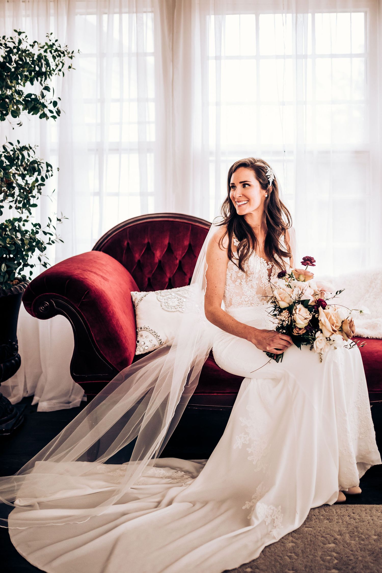 Elegant bride in flowing white gown seated on red velvet couch with floral bouquet.